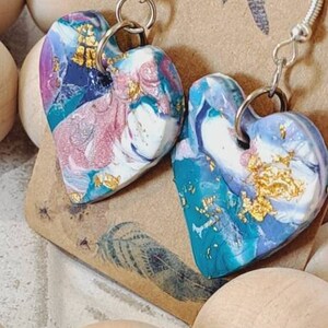 May include: A pair of heart-shaped earrings with a blue, white and gold marbled design. The earrings are made of polymer clay and have silver hooks.