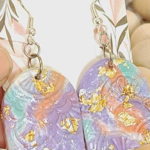 May include: A pair of handmade earrings with a purple, pink and green marbled design. The earrings have gold flakes embedded in the resin and are shaped like arches. The earrings are hanging from silver hooks.