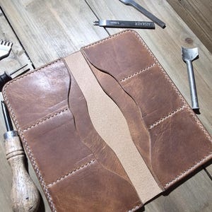 Puede incluir: Cartera de cuero marrón hecha a mano con múltiples ranuras para tarjetas y un bolsillo central. La cartera está abierta, revelando el diseño interior. Herramientas de marroquinería dispuestas alrededor de la cartera.