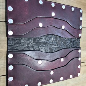Puede incluir: Una cartera de cuero burdeos con un panel central decorativo tallado. La cartera presenta un diseño en capas con detalles de puntos blancos a lo largo de los bordes. El diseño general es rectangular y parece hecho a mano.