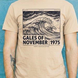 Puede incluir: Una camiseta de manga corta color canela con un gráfico en tinta negra de olas rompiendo bajo un cielo tormentoso. El texto dice "GALES OF NOVEMBER 1975" con "Lake Superior" y coordenadas sobre la ilustración, y una línea de texto.