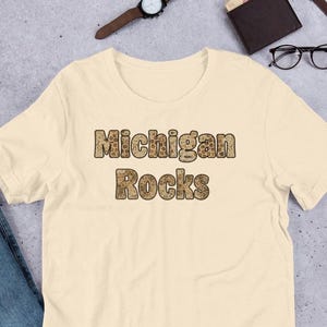 May include: A cream-colored short-sleeve t-shirt featuring the text Michigan Rocks printed in a brown and tan leopard print pattern across the center of the chest. The shirt has a crew neckline and a standard fit with short sleeves and a straight hem.