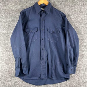 May include: A long-sleeve navy blue button-down shirt featuring a pointed collar, a central button placket, and two chest pockets with buttoned flaps. The shirt has matching navy blue buttons and buttoned cuffs on each long sleeve.
