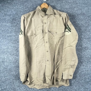 May include: A long-sleeve tan button-down shirt featuring two front chest pockets with button flaps. The sleeves display military-style patches with dark green chevron stripes and crossed rifle insignias on a light tan circular background.