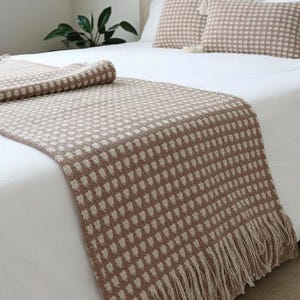 Könnte beinhalten: Ein gewebter Bettläufer und passende Kissen in einer neutralen Farbpalette. Der Läufer weist ein sich wiederholendes Muster aus kleinen Quadraten in Beige und Off-White auf, mit einem Fransenrand. Die Kissen haben ein ähnliches Design und Quasten.