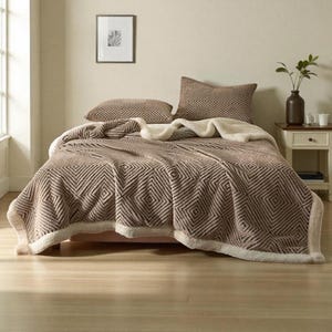Pode incluir: Um conjunto de cama castanho e creme com um padrão geométrico de losangos. O conjunto inclui um cobertor e duas almofadas. O cobertor tem uma textura macia e felpuda com uma borda creme. As almofadas têm um padrão de losangos a condizer.