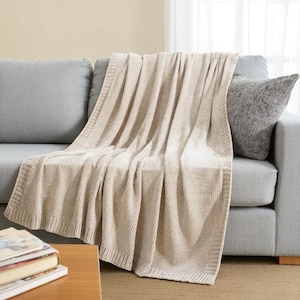 May include: A beige knitted throw blanket draped over a gray sofa. The blanket has a textured, waffle-like pattern and a ribbed border. A gray pillow is visible on the sofa. The blanket is made of a soft, comfortable material.