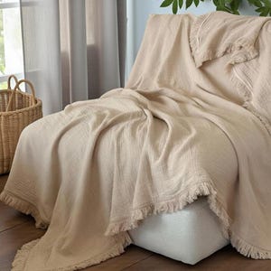 Puede incluir: Una manta de algodón beige con borde de flecos está drapeada sobre una silla blanca. La manta tiene un aspecto texturizado y está doblada para crear un efecto de capas. Una cesta de mimbre es visible en el fondo.