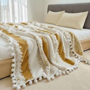 Puede incluir: Una manta suave, blanca y dorada con un patrón texturizado y ribete de pompones blancos. La manta está sobre una cama con almohadas color crema y una cabecera gris. El diseño de la manta presenta una franja dorada.
