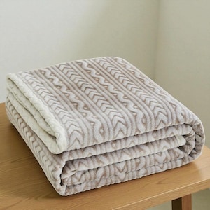May include: Folded beige and white blanket with a textured, patterned design. The blanket features a repeating arrow-like pattern in a lighter beige color against a darker beige background. The edges are neatly finished, and the blanket is folded in thirds.