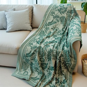 May include: A green and white floral patterned throw blanket draped over a beige sofa. The blanket features a detailed botanical design with a border. Two striped cushions are visible on the sofa. The blanket is made of a soft material.