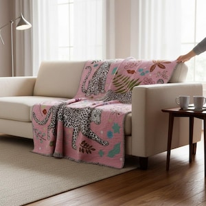 Puede incluir: Una manta rosa con un diseño de estampado de leopardo, con manchas negras sobre un fondo blanco. La manta también tiene patrones florales y de hojas en azul, verde y rosa. La manta está sobre un sofá beige.