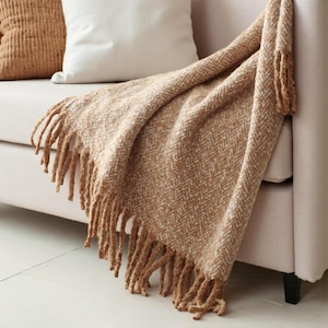 Pode incluir: Uma manta tecida bege e creme com franjas, drapeada sobre um sofá bege. A manta tem um padrão de espinha de peixe texturizado. Duas almofadas são visíveis atrás da manta.