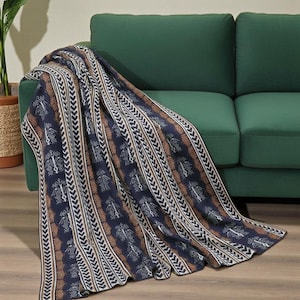 May include: A navy blue and tan patterned throw blanket draped over a green sofa. The blanket features a repeating geometric design with white, tan, and navy blue elements. The throw blanket is made of a soft material.