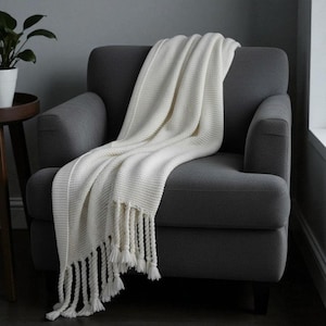 May include: A cream-coloured knitted throw blanket with fringe detail draped over a grey armchair. The blanket has a textured weave and a soft appearance. The tassels hang down the side of the chair.