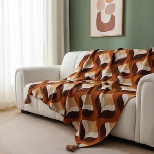 Peut inclure: Une couverture à motifs marron, beige et blanche, avec des pompons, drapée sur un canapé blanc. La couverture présente un motif géométrique avec des lignes courbes, lui donnant un aspect rétro. Les pompons sont de couleur marron.
