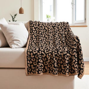 May include: A black and tan leopard print blanket is draped over a beige sofa. The blanket has a repeating animal spot pattern and a tan border. Two white cushions are visible on the sofa.