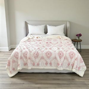 May include: A cosy blanket with a geometric pattern in white and pale pink, draped over a bed with white pillows. The blanket has a sherpa-like texture and a warm appearance, ideal for adding comfort and style to a bedroom.
