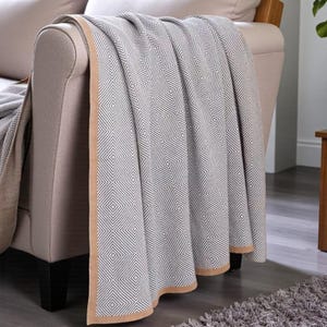 May include: A grey and white geometric patterned throw blanket draped over a beige sofa. The blanket features a diamond pattern and a tan border. The sofa has wooden legs and is placed on a grey rug. The blanket is a home decor item.