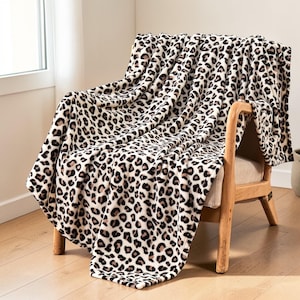 May include: A soft, plush blanket with a leopard print design draped over a wooden chair. The blanket features a cream-colored background with brown and black spots. The chair has a light beige seat and a natural wood frame.