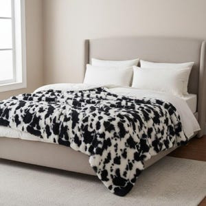 Puede incluir: Una manta con estampado en blanco y negro sobre una cama. La manta tiene un diseño de manchas similar al de una vaca. La cama tiene almohadas blancas y un cabecero de color neutro. La habitación tiene una combinación de colores neutros.