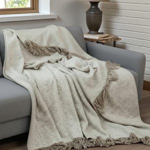 May include: A neutral-toned throw blanket with fringe detail draped over a grey sofa. The blanket has a textured weave and a soft appearance. The fringe is a darker shade of brown.