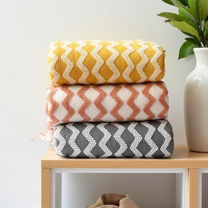 May include: Three decorative cushions with a zig-zag pattern in shades of yellow, peach, and grey. The cushions are stacked on a wooden shelf. The top cushion is yellow, the middle is peach, and the bottom is grey.