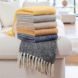 May include: A selection of folded blankets in shades of yellow, grey, and navy, featuring a herringbone design. The bottom blanket is black and white with a fringed edge.