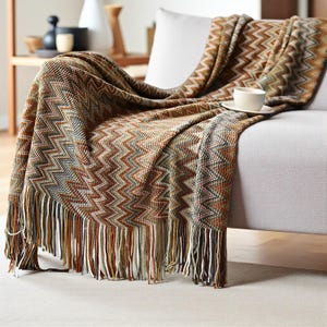 Könnte beinhalten: Eine gewebte Decke mit Zickzackmuster in Braun-, Grün- und Orangetönen, drapiert über einem grauen Sofa. Die Decke hat einen Fransenbesatz am unteren Rand. Eine weiße Tasse und Untertasse liegen darauf.