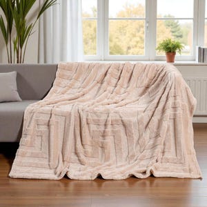 Könnte beinhalten: Eine beige Kuscheldecke mit geometrischem Muster, drapiert über einem grauen Sofa. Die Decke hat eine weiche Textur. Im Hintergrund sind ein Fenster und eine Topfpflanze zu sehen. Die Decke ist wahrscheinlich aus weichem Material.