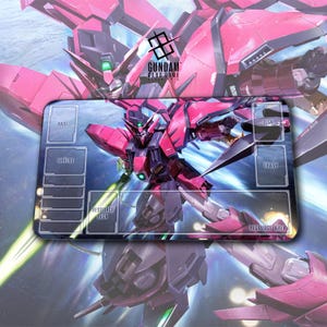 May include: A rectangular Gundam card game playmat featuring a red and black robot design. The playmat has labeled zones for base, shields, resource deck, deck, trash, and resource area. The Gundam logo is in the upper center.