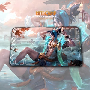 Puede incluir: Alfombrilla rectangular para cartas de juego Riftbound League of Legends. Presenta una ilustración detallada de un personaje con atuendo azul y dorado, sobre un fondo de agua y hojas de otoño. La alfombrilla tiene zonas numeradas para colocar las cartas y el texto "Riftbound League of Legends Trading Card Game".
