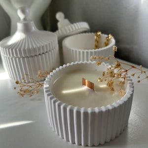 Peut inclure: Un bougeoir en céramique blanche côtelée avec une mèche en bois, rempli de cire. D'autres récipients en céramique blanche et des fleurs séchées sont également visibles. Thème : décoration intérieure.
