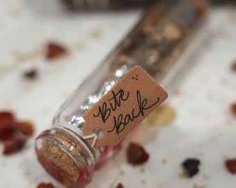 May include: A clear glass vial filled with small dark botanical pieces and silver metallic flakes, sealed with a cork stopper. A small brown paper tag is tied to the neck with string, featuring the handwritten black ink text Bite Back.