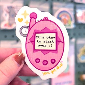 May include: A pink sticker featuring a retro handheld game design. The screen displays the text "It's okay to start over :)". Additional elements include yellow stars, a heart, and the phrase "You got this!" in a script font.