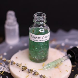 May include: A clear glass bottle filled with green liquid and small particles, labeled "Religious Trauma Healing Intention Oil." A dropper with green liquid is next to the bottle. The bottle sits on a white, round surface with lavender sprigs.