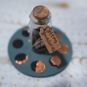 May include: A clear glass bottle with a cork stopper sealed with white wax, containing dark contents. A brown paper tag attached with string features the handwritten text "Setting Boundaries". The bottle sits on a circular blue base with copper-colored moon phase accents.