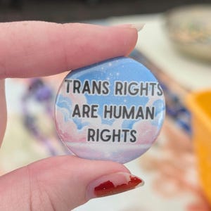 Może przedstawiać: Okrągły przypinany znaczek z tłem przedstawiającym jasnoniebieskie i różowe niebo z białymi chmurami. Napis "TRANS RIGHTS ARE HUMAN RIGHTS" jest wydrukowany pogrubionymi czarnymi wielkimi literami na środku powierzchni znaczka.