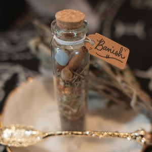 May include: A clear glass vial with a cork stopper, filled with a mixture of dried herbs, small stones, and a cinnamon stick. A small brown paper tag tied with white string features the handwritten word Banish in black ink.