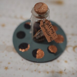 May include: A small clear glass bottle with a cork stopper sealed with white wax, containing dark contents. A brown paper tag tied to the neck reads Setting Boundaries. The bottle sits on a circular teal base featuring painted moon phases.