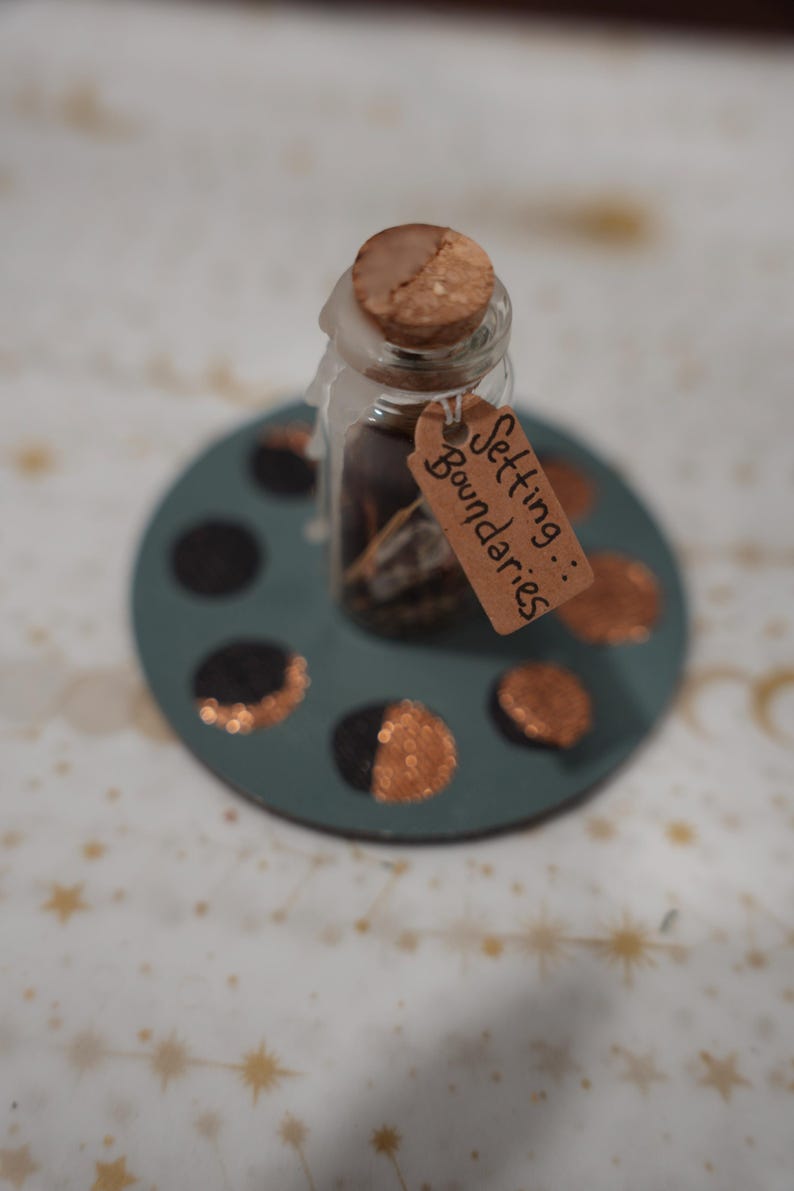 May include: A small clear glass bottle with a cork stopper sealed with white wax, containing dark contents. A brown paper tag tied to the neck reads Setting Boundaries. The bottle sits on a circular teal base featuring painted moon phases.