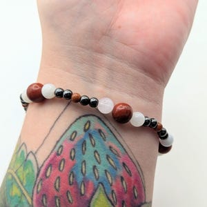 May include: A beaded bracelet featuring a repeating pattern of large round reddish-brown stones, smaller white translucent beads, metallic dark grey spherical beads, and tiny brown accent beads arranged on a flexible cord around the wrist.