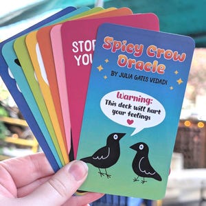 May include: A hand holding a fan of colorful oracle cards. The top card reads "Spicy Crow Oracle" with the author's name and a warning about hurting feelings. The cards feature a crow illustration and various colors.