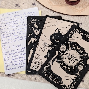 May include: A collection of tarot cards featuring black and white cat illustrations. One card reads "TAROT" in a circular design. A handwritten note with card interpretations is also present.