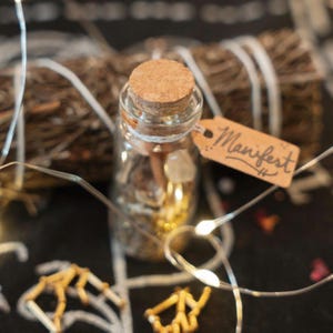 May include: A clear glass bottle with a cork stopper, containing small crystals and gold-colored elements. A small brown paper tag tied to the neck features the handwritten word Manifest. A bundle of dried twigs is positioned behind the bottle.