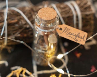May include: A clear glass bottle with a cork stopper containing small crystals and gold-toned items. A small brown paper tag tied to the neck with white string features the handwritten word Manifest. A bundle of dried twigs is visible behind.