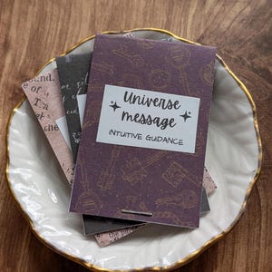 May include: A small stack of cards with the text "Universe message" and "Intuitive Guidance" on a purple background with key illustrations. The cards are in a white dish with a gold rim, set on a wooden surface.