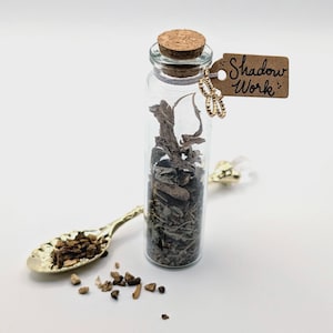 May include: A clear glass bottle with a cork stopper, filled with dried herbs, and a small tag that reads "Shadow Work". A gold-colored spoon is next to the bottle, with herbs in the spoon and scattered around it.