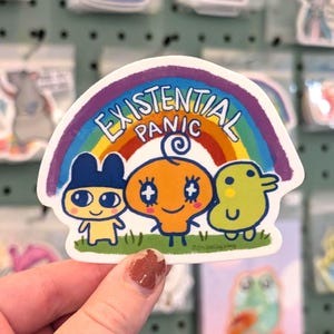 May include: A white sticker featuring three cartoon characters under a rainbow with the words "EXISTENTIAL PANIC". The characters are yellow, orange, and green, standing on a green grassy area. The rainbow has purple, blue, green, yellow, and red stripes.