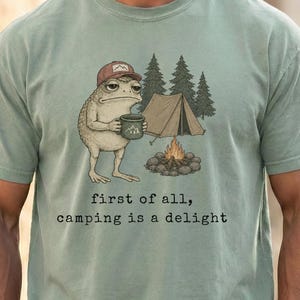 May include: A sage green t-shirt featuring a graphic of a toad wearing a baseball cap and holding a mug. The illustration includes a tent, pine trees, and a campfire. Below the graphic, the text reads: first of all, camping is a delight.
